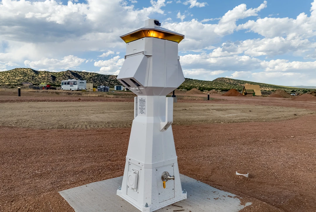 Close-up of white electrical pedestal for RV hookups featuring multiple outlet panels and utility connections, installed on concrete pad at campground construction site with rolling hills and blue sky in background