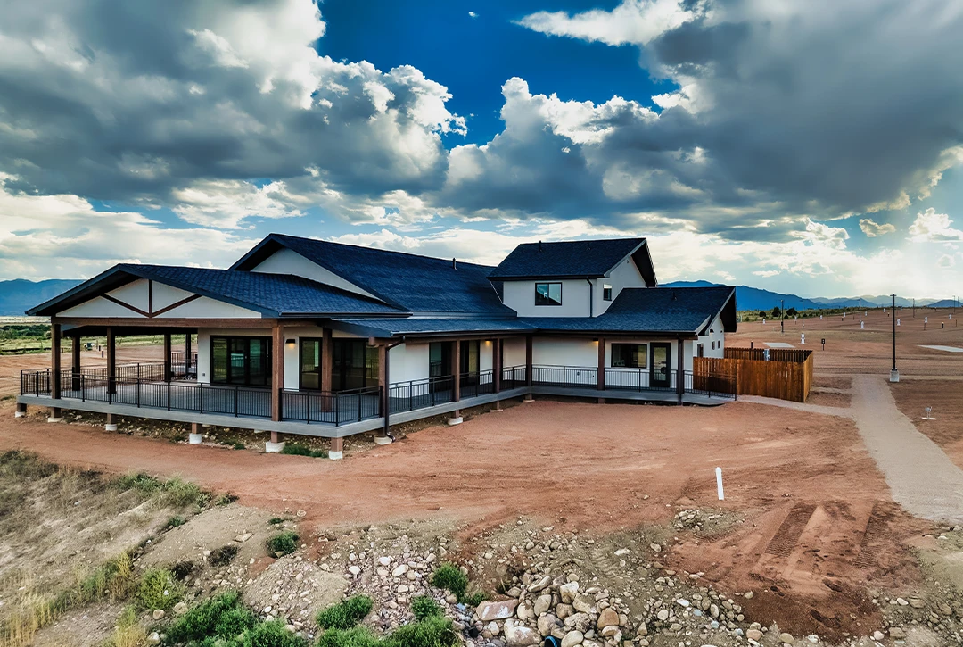 Newly constructed Observer Ranch Campground main building featuring modern architecture with dark blue metal roofing, extensive covered wraparound deck with metal railings, large windows, and stone accents, set against dramatic cloudy sky and mountains
