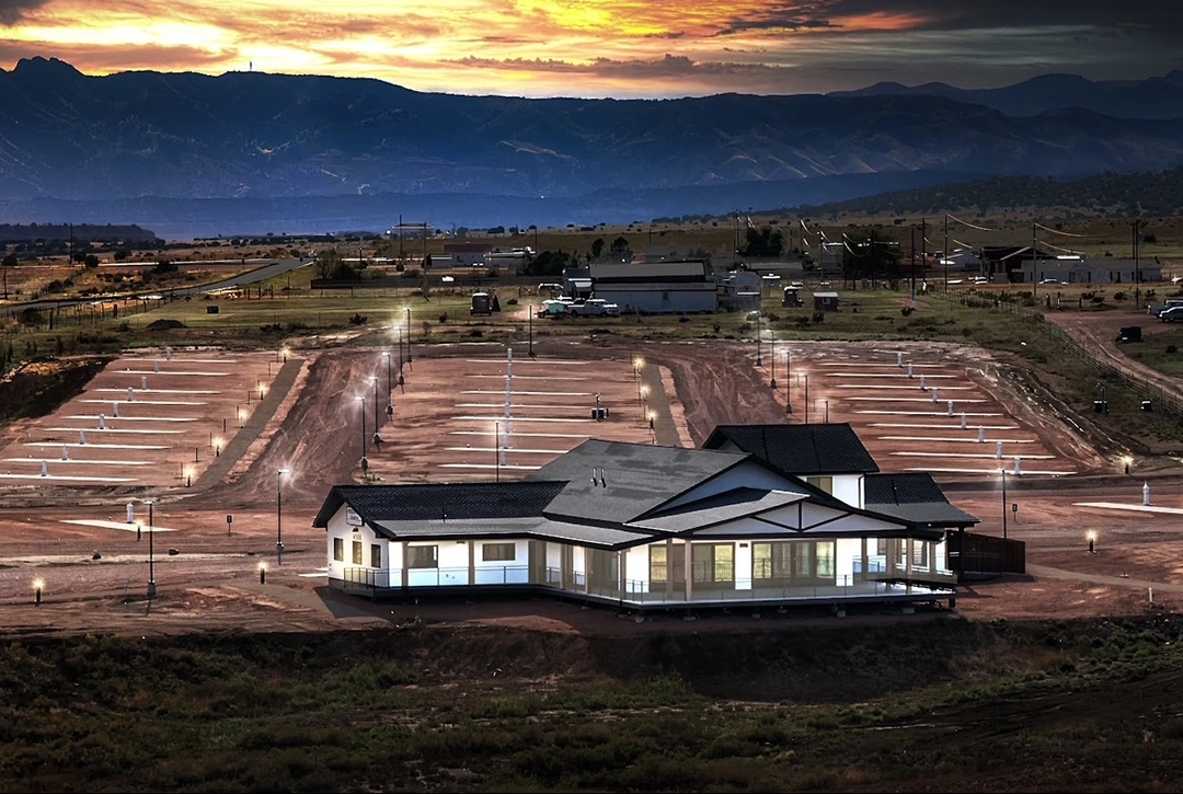 Aerial view of a completed RV campground at sunset showing a modern white ranch-style building with dark roofing in the center, surrounded by organized RV sites with electrical hookup pedestals and gravel pads, set against Colorado mountains with dramatic