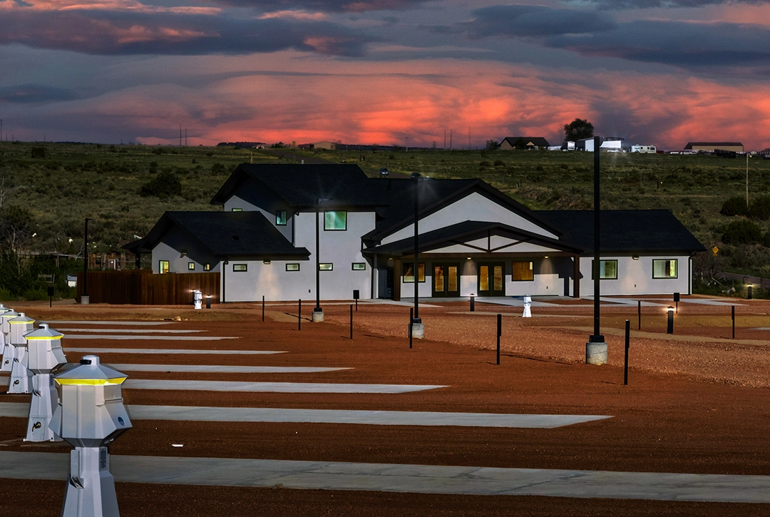Modern campground facilities building photographed at dusk with dramatic pink and orange sunset clouds overhead, featuring white exterior walls, dark roofing, covered entry portico, and organized parking area with electrical pedestals for RV hookups in th