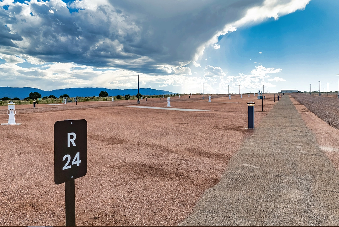 RV campground site layout showing numbered site R24 in foreground with black and white signage, gravel camping pads, white electrical hookup pedestals arranged in rows, and Colorado mountains visible in the background under a dramatic cloudy sky