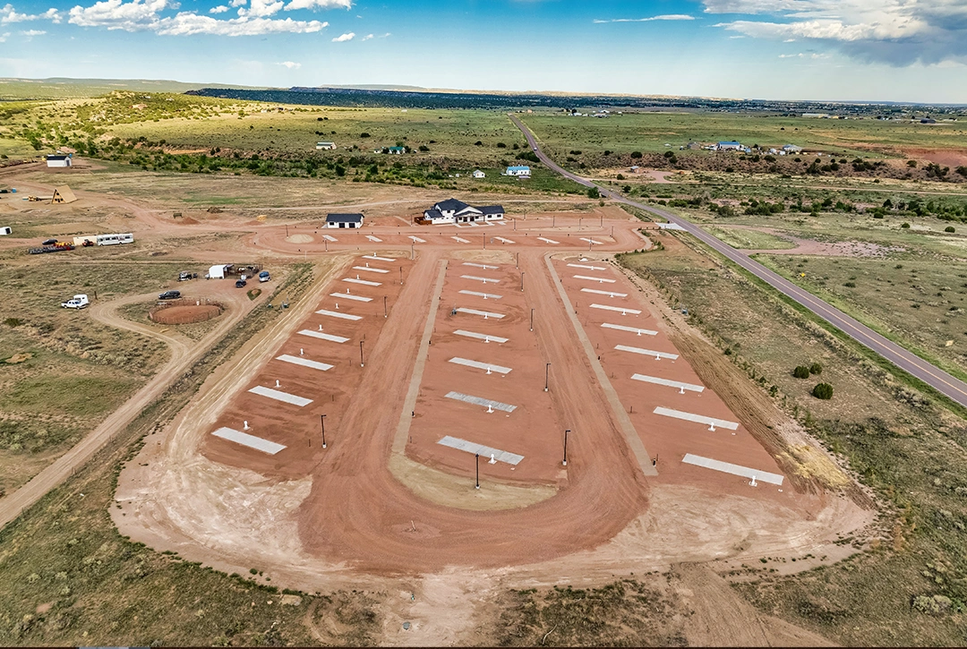 Aerial view of a completed RV campground development in rural Colorado showing organized rows of white concrete RV pads with electrical hookup pedestals on reddish dirt terrain, featuring a modern facilities building with dark roofing at the top of the la